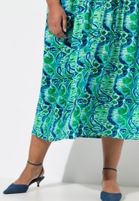 Grün- und blau gemustertes Kleid mit fließendem Design, knielang. Getragen mit blauen Slingback-Pumps mit spitzem Zehenbereich und niedrigem Absatz.