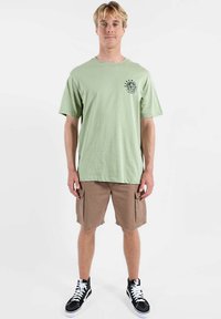 T-shirt en coton vert clair avec un col rond et un logo circulaire noir sur la poitrine, associé à un short cargo marron et des baskets noires.