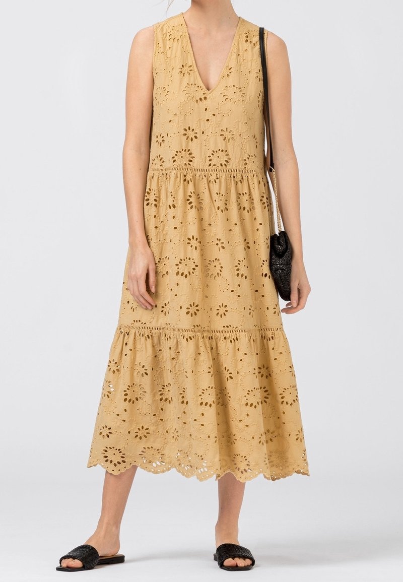 Vestido sin mangas en caqui con diseño de ojales florales, escote en V, cintura fruncida y falda en capas. Combinado con sandalias negras tipo slip-on y un bolso pequeño.