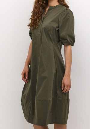 Femme portant une robe ample vert olive longueur genoux avec des manches courtes bouffantes et une fente en V, debout devant un fond clair uni.