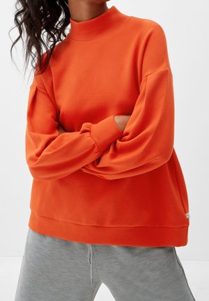 Person wearing bright orange long-sleeve pullover with high collar and loose gray pants, arms crossed, against plain white background.