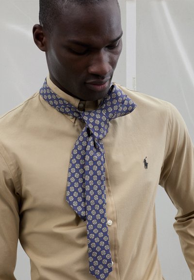 Sciarpa blu con motivo a esagoni indossata sopra una camicia beige con bottoni, dotata di colletto e di un piccolo logo sul petto a sinistra.