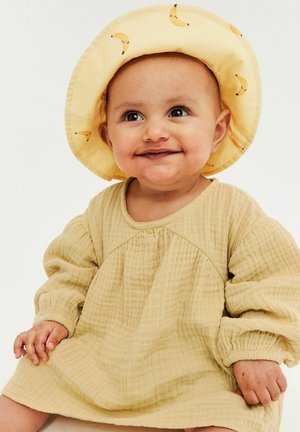 Lächelndes Baby, das ein gelbes strukturiertes Kleid und einen gelben Hut mit kleinen Bananenmotiven trägt, sitzt vor einem schlichten hellen Hintergrund.