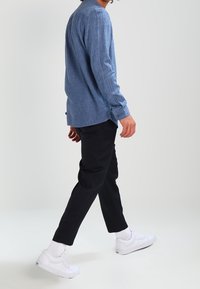 Camicia a maniche lunghe in chambray azzurro chiaro, vestibilità rilassata, indossata con pantaloni neri a sigaretta e sneakers bianche, presentando un outfit informale.