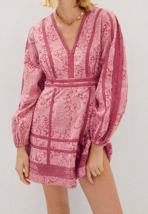 Femme portant une mini-robe rose à motifs, manches longues, avec un décolleté en V profond et un collier doré décoratif.