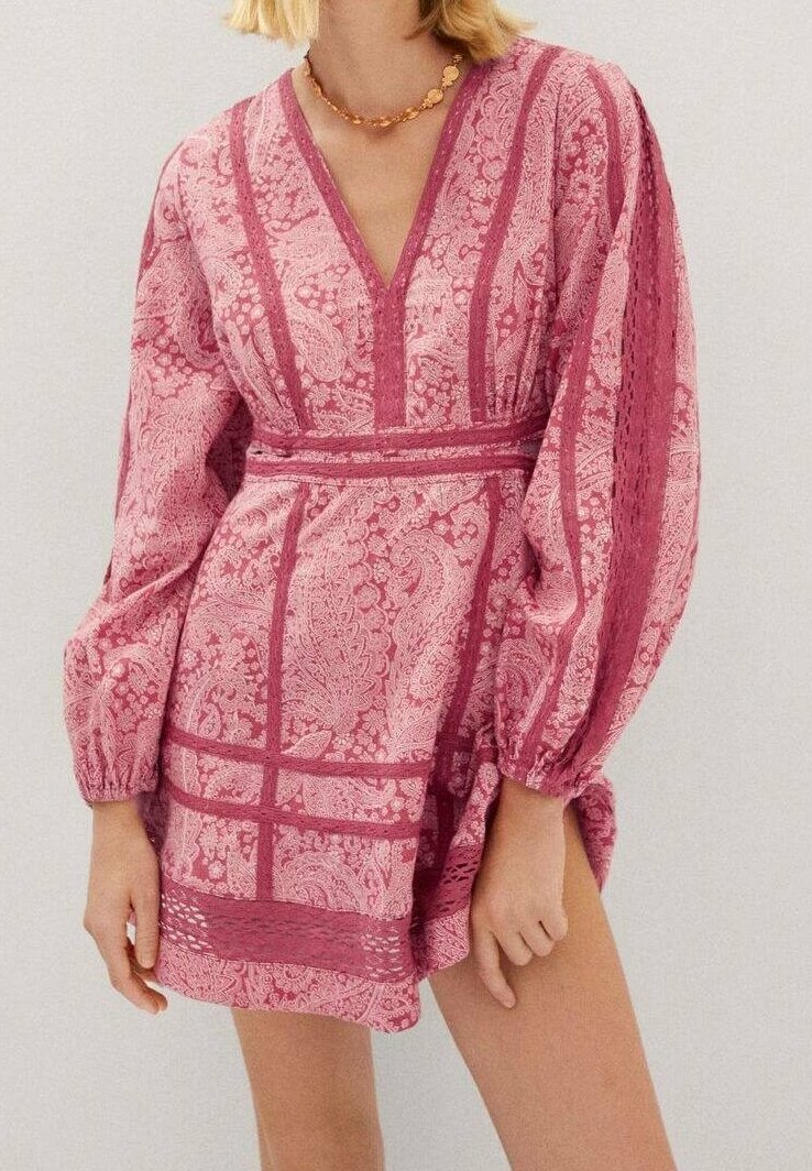 Femme portant une mini-robe rose à motifs, manches longues, avec un décolleté en V profond et un collier doré décoratif.