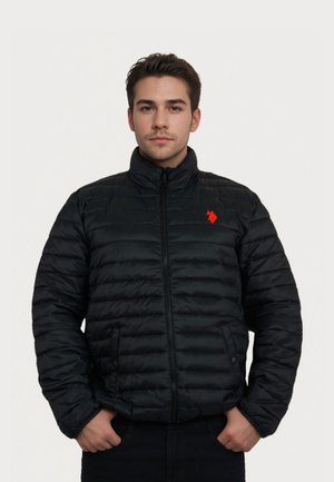 Junger Mann trägt eine schwarze gesteppte Jacke mit rotem Logo und steht mit den Händen in den Taschen vor einem einfarbigen hellen Hintergrund.