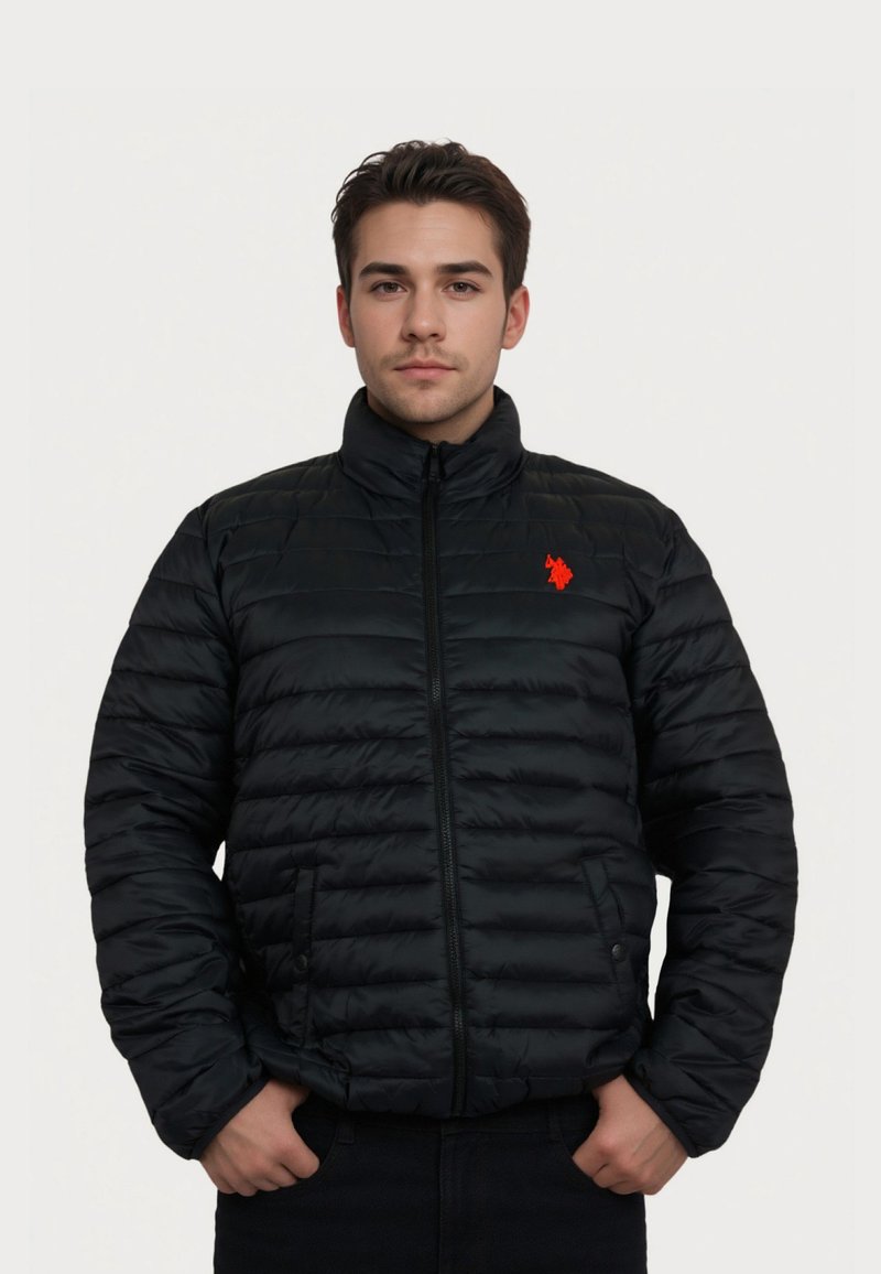 Junger Mann trägt eine schwarze gesteppte Jacke mit rotem Logo und steht mit den Händen in den Taschen vor einem einfarbigen hellen Hintergrund.