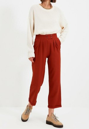 Frau trägt einen cremefarbenen Zopfstrickpullover, hoch taillierte rostrote Hose mit Knöpfen, aufgerollten Bündchen und beigefarbenen Schnürschuhen.