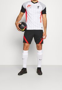 Nike Liverpoolin jalkapallopaita, jossa on harmaa raidallinen paita punaisilla yksityiskohdilla, mustat shortsit punaisilla yksityiskohdilla, valkoiset sukat ja mustat jalkineet.
