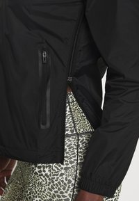 Sort agil jakke med lynlåslomme og elastiske manchetter. Kombineret med grønne leggings med leopardmønster for et kontrastfuldt design.