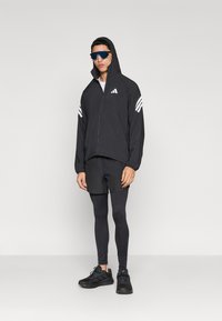 Zwarte sportjas met capuchon, witte streepdetails, gecombineerd met zwarte korte broek en legging. Model draagt zwarte sneakers en een zonnebril.