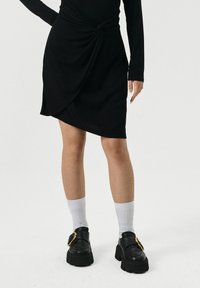 Vrouw in een zwarte asymmetrische rok, een langarmige top, witte sokken en zwarte stevige loafers met een grote gouden gesp.