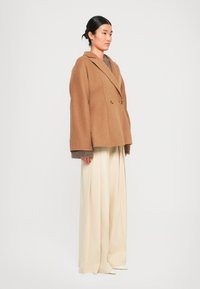 Manteau double boutonnage en laine marron avec des manches larges, associé à un pull gris et un pantalon large crème. Textures lisses et boutons dorés.