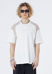 White t-shirt with light beige raglan sleeves, crew neck, and subtle logo at center. Features relaxed fit and soft fabric texture.
