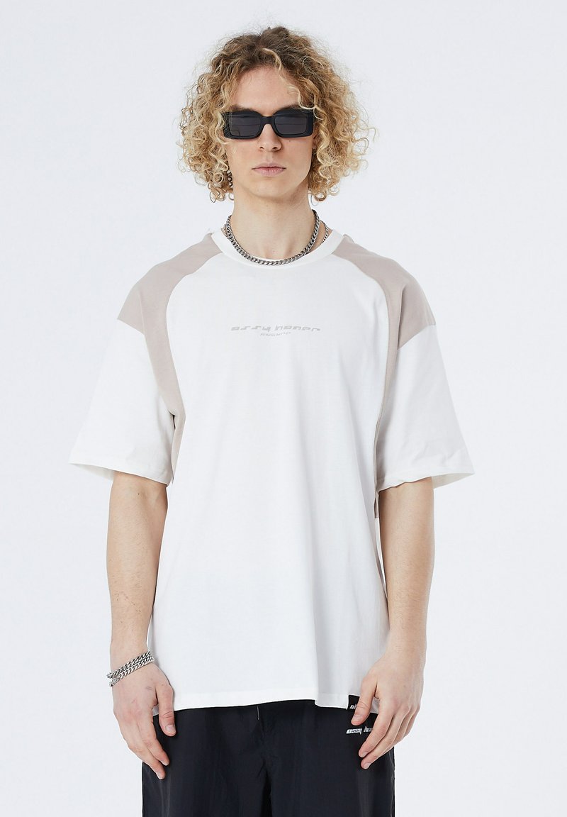 White t-shirt with light beige raglan sleeves, crew neck, and subtle logo at center. Features relaxed fit and soft fabric texture.