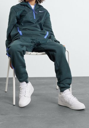 Osoba ubrana w ciemnozielony dres Nike i białe buty sportowe Nike, siedząca na białym krześle na jednolitym tle.