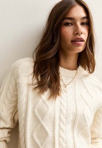 Kremowy sweter w warkocze z wzorem w diamenty i skręconymi liniami. Miękka tekstura, okrągły dekolt, a na nim delikatny złoty naszyjnik.