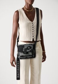 Black and grey patterned crossbody bag with a flap closure, featuring a logo and adjustable strap, worn with a textured white top and light pants.