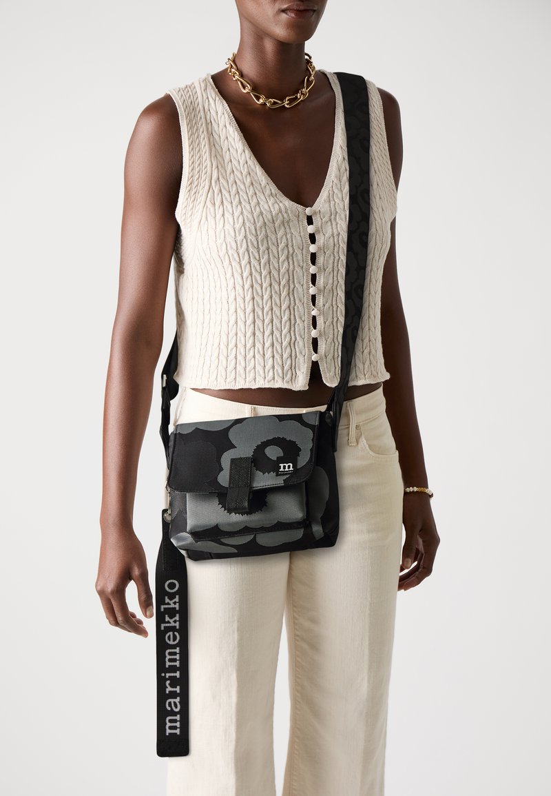 Black and grey patterned crossbody bag with a flap closure, featuring a logo and adjustable strap, worn with a textured white top and light pants.