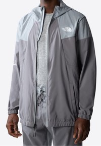 Harmaa tuulihaalari, jossa on vaaleanharmaat yksityiskohdat, säädettävä nyörimäinen helma, vetoketjuluukku ja logo rinnassa. Sujuva pinta, kevyt muotoilu.