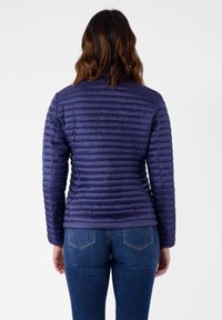Giacca imbottita viola con design trapuntato e chiusura con zip, caratterizzata da un colletto eretto e maniche lunghe. Indossata con jeans blu.