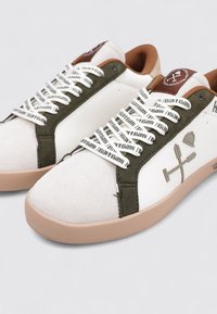 Zapatillas deportivas blancas con detalles en ante verde, con detalles marrones, cordones con patrón y suela de goma. Logo notable de la marca en el lateral.