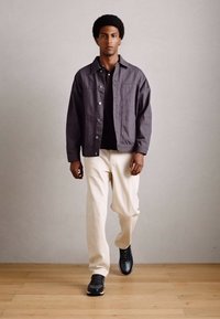 Mann, der drinnen auf einem Holzboden geht, trägt eine dunkelgraue Jacke, ein schwarzes Hemd, cremefarbene Hosen und schwarze Sneaker.