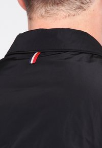 Černá bunda se sklopeným límcem a malou trojbarevnou cedulkou (červená, bílá, modrá) na zadní straně límce. Hladká textura a lehký materiál.