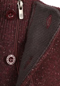 Bordeauxrode gebreide trui met rits, voorzien van een donkergrijze voering en een knoopdetail. Textuurstof met spikkels is zichtbaar.
