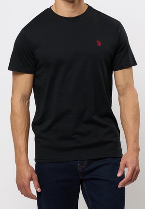 Mann trägt ein schlichtes schwarzes T-Shirt mit kurzen Ärmeln und einem kleinen roten gestickten Logo auf der linken Brust, kombiniert mit dunkelblauen Jeans.