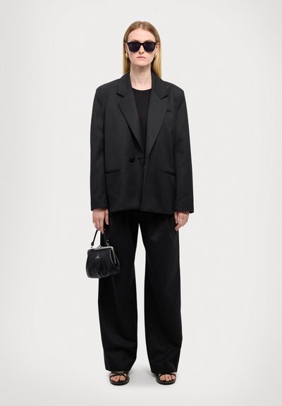 Svart oversized blazer med spiss krage, parret med en svart topp og brede bukser. Tilbehøret inkluderer svarte solbriller og en liten håndveske.
