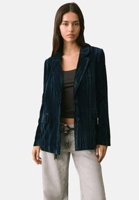 Blazer din catifea bleumarin cu rever decupat, două butoane și buzunare laterale. Textură netedă și croială lejeră; asortat cu o bluză neagră și pantaloni gri deschis.