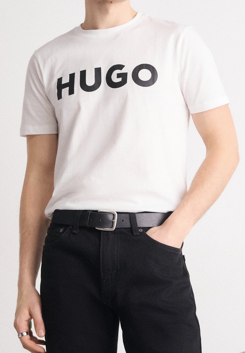 Biała bawełniana koszulka z dużym czarnym napisem "HUGO". Noszona z czarnymi dżinsami i czarnym paskiem. Minimalistyczny design, swobodny krój.