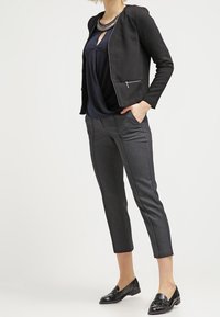 Schwarze Blazer mit einer Reißverschlusstasche, über einer marineblauen Bluse getragen. Graue maßgeschneiderte Hose mit schmalem Schnitt und schwarzen Loafers. Glatte Textur insgesamt.