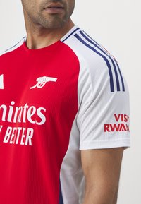 Maillot de football rouge et blanc en tissu mesh, avec un logo sur la poitrine et le texte "Emirates" et "Visit Rwanda" sur les manches.
