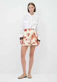 Witte overhemd met knopen en twee borstzakken, gecombineerd met crèmewitte shorts met een oranje bloemenpatroon. Simpele platte schoenen maken de look compleet.