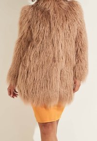 Mujer vestida con un abrigo largo de piel sintética marrón claro sobre una falda naranja brillante hasta la rodilla, de pie frente a un fondo sencillo.
