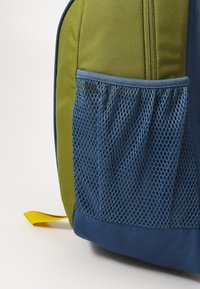Σακίδιο με πράσινο υφασμάτινο σώμα, μπλε πλευρική τσέπη από δίχτυ και κίτρινη λαβή. Διαθέτει ανθεκτική ραφή και δομημένο σχήμα.