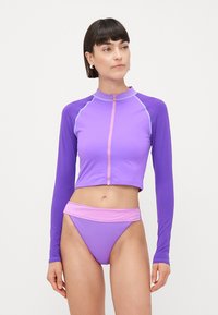 Violets garš rukavu peldkostīms ar rāvējslēdzēju un kontrastējošām gaiši violetām akcentiem, papildināts ar atbilstošām augsto jostasvietu bikini apakšdaļām.