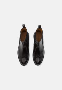 Bottes Chelsea en cuir noir poli avec détails brogue et languettes de traction, vues de dessus sur un fond clair uni.