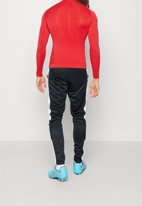 Czerwona sportowa koszulka z długim rękawem w połączeniu z czarnymi spodenkami dresowymi z białymi bocznymi paskami, noszona z niebieskimi korkami piłkarskimi.