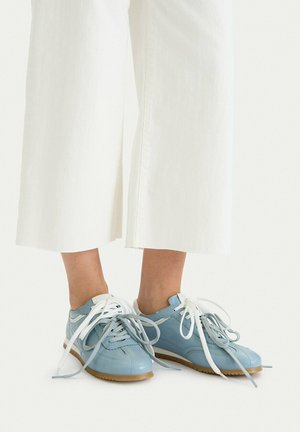 Person trägt hellblaue Ledersneaker mit weißen und grauen Schnürsenkeln, kombiniert mit weiten weißen Hosen, die oberhalb der Knöchel abgeschnitten sind.