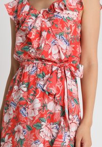 Červené květinové šaty s volány na ramenech, zdobené růžovými a modrými květy, s páskem v pase a lehkým, splývavým texturem.