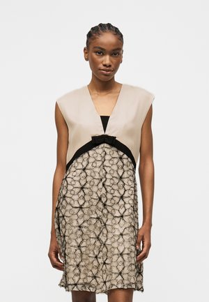 Kobieta w beżowej sukience bez rękawów z głębokim dekoltem w kształcie litery V, czarną kokardką i teksturowaną czarną koronkową nakładką na spódnicy.