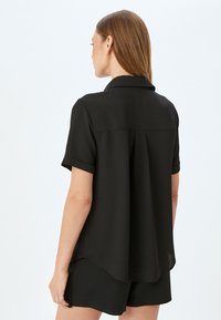 Blusa negra de manga corta con cuello puntiagudo, tejido suave, detalle de pliegues en la parte posterior y dobladillo redondeado, combinada con pantalones cortos negros a juego.