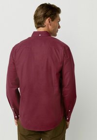 Camisa de botones de manga larga color burdeos con cuello, que presenta una textura suave y un brillo sutil. Tiene un dobladillo redondeado y puños abotonados.