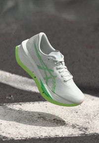 Baskets blanches avec des accents verts, tige perforée, bout arrondi et semelle légère, conçues pour la course ou les activités sportives.