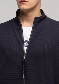 Granatowa kurtka zapinana na zamek z teksturowanej tkaniny, wysoki kołnierz oraz kontrastująca biała koszulka pod spodem z niebieską grafiką.