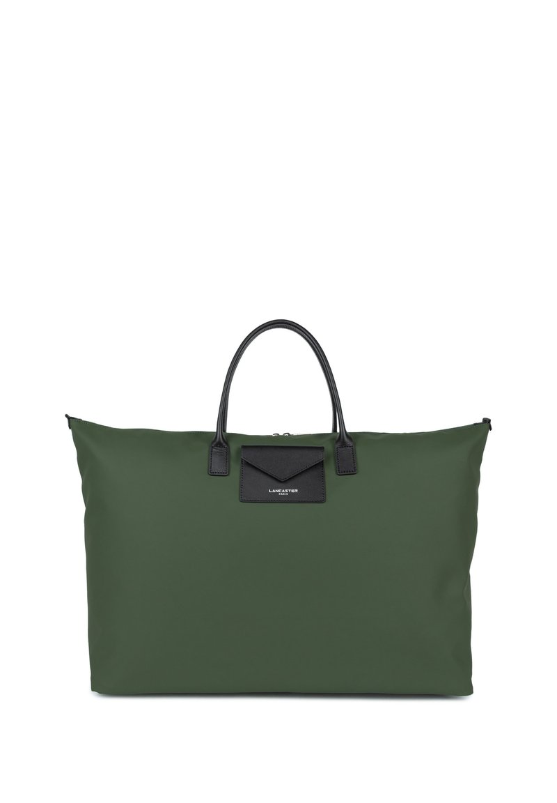 Green fabric tote bag with black leather accents, structured shape, dual handles, and a black patch with logo detail on the front.